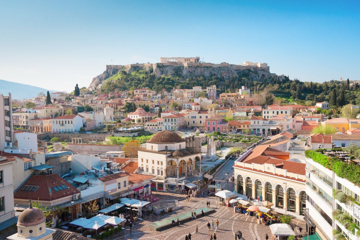 An elevated view of a bustling square in Athens, with colorful buildings and a view of the Acropolis in the distance.